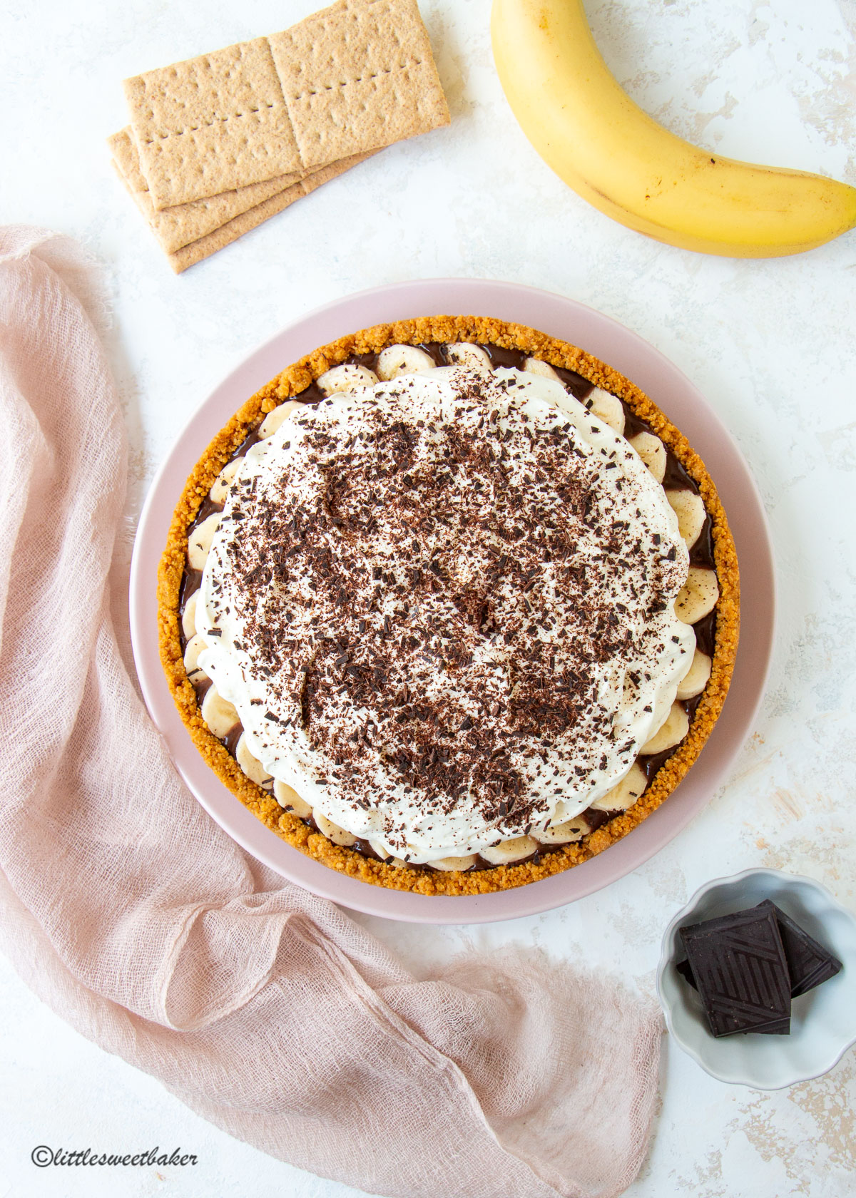 A chocolate banoffee pie surrounded by a banana, graham crackers, dark chocolate in a bowl and a pink napkin.