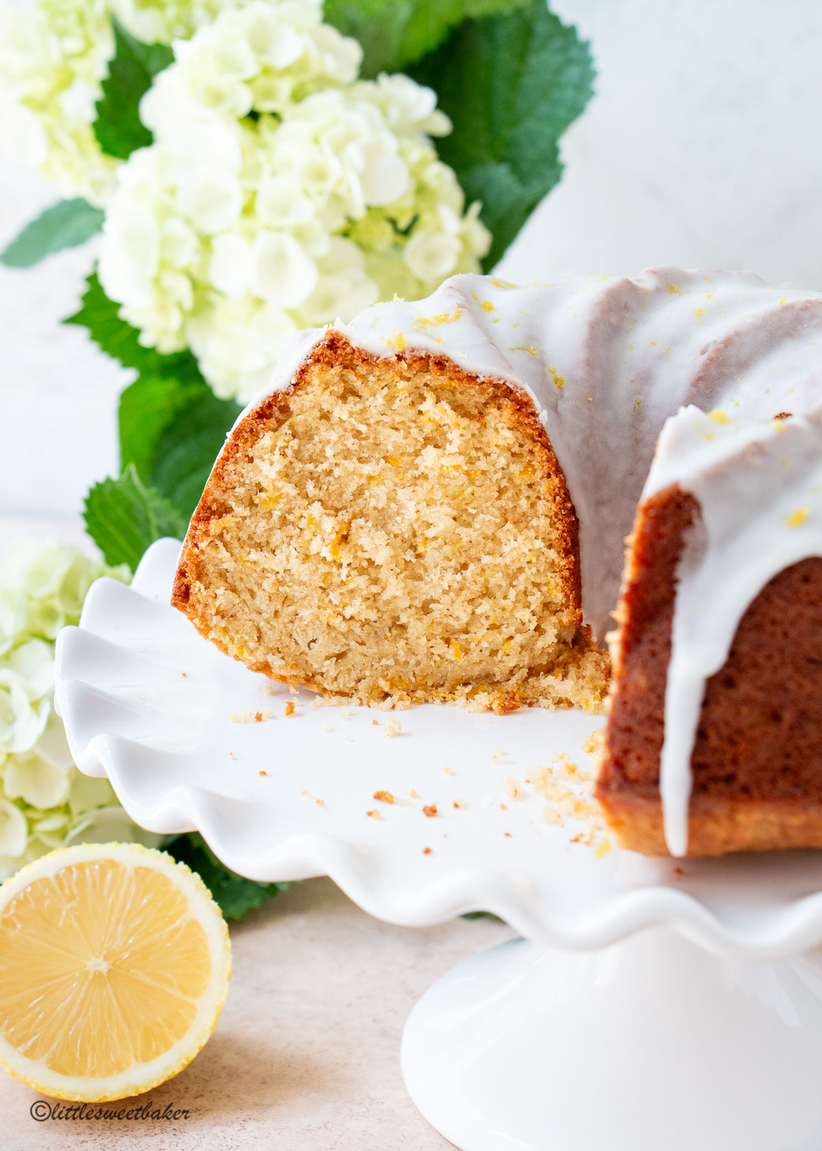 A lemon zucchini cake on a white cake stand with white flowers in the back.