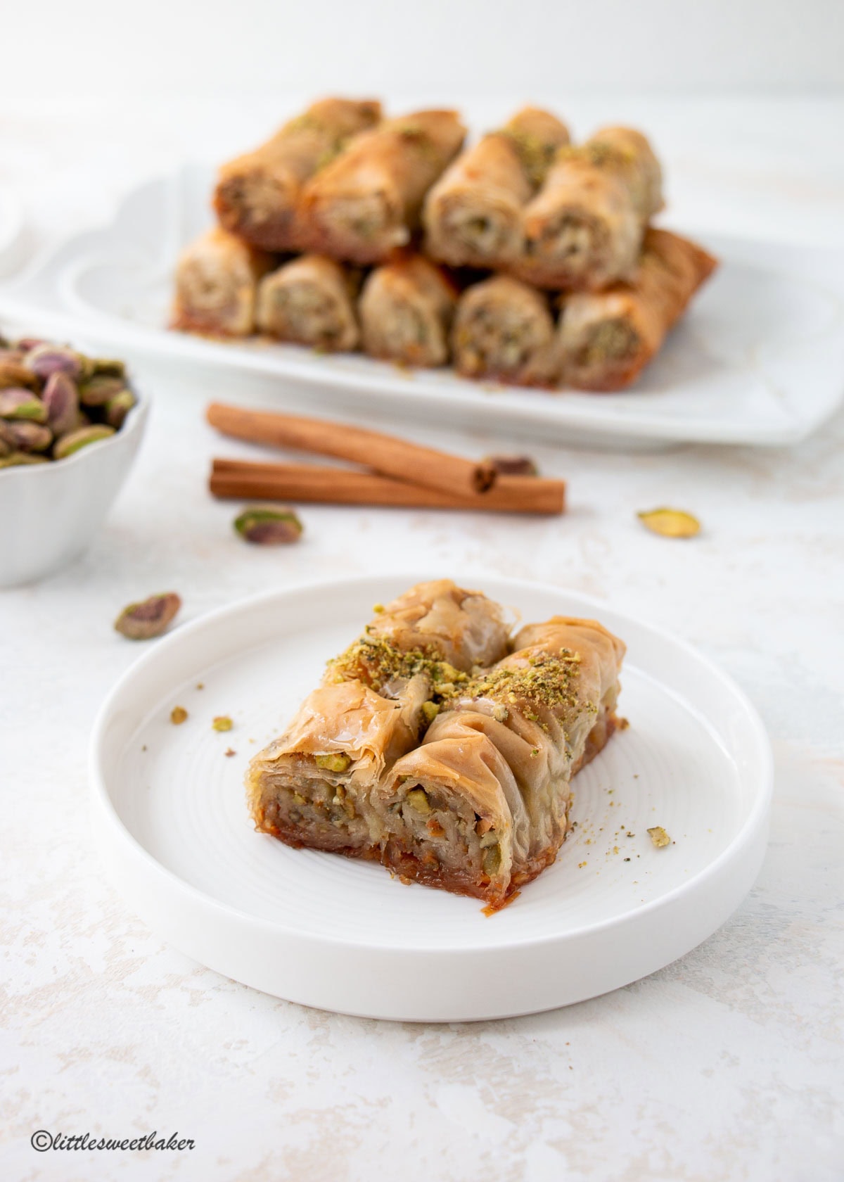 Two pistachio baklava rolls on a white plate with a bowl of pistachios and a serving plate of rolls.