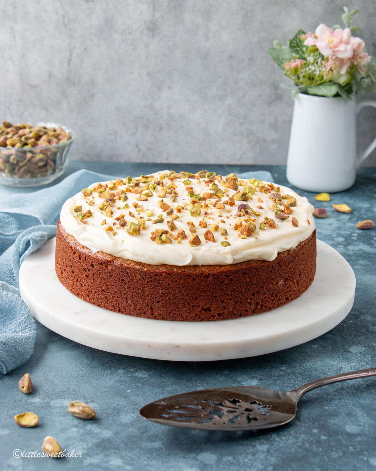 A pistachio cake topped with cream cheese frosting on a marble cake platter.