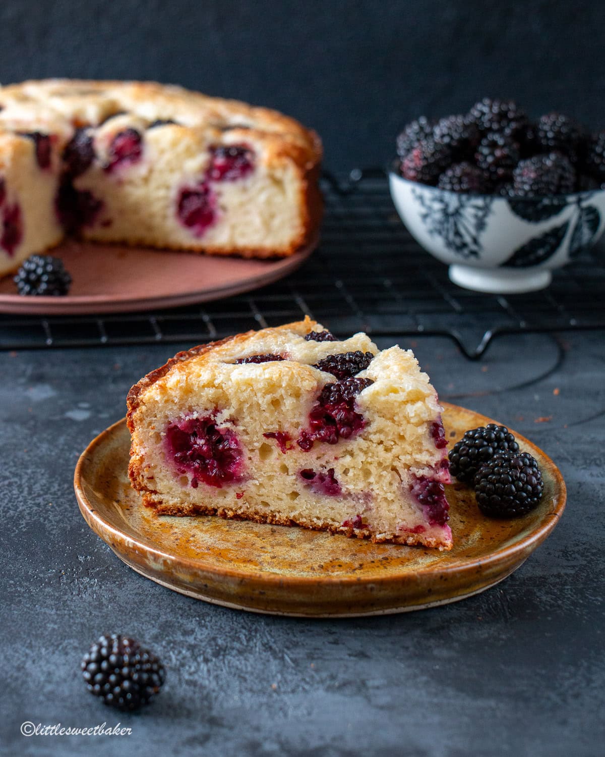 A slice of blackberry cake on a pottery plate with the rest of the cake and a bowl of blackberries in the background.