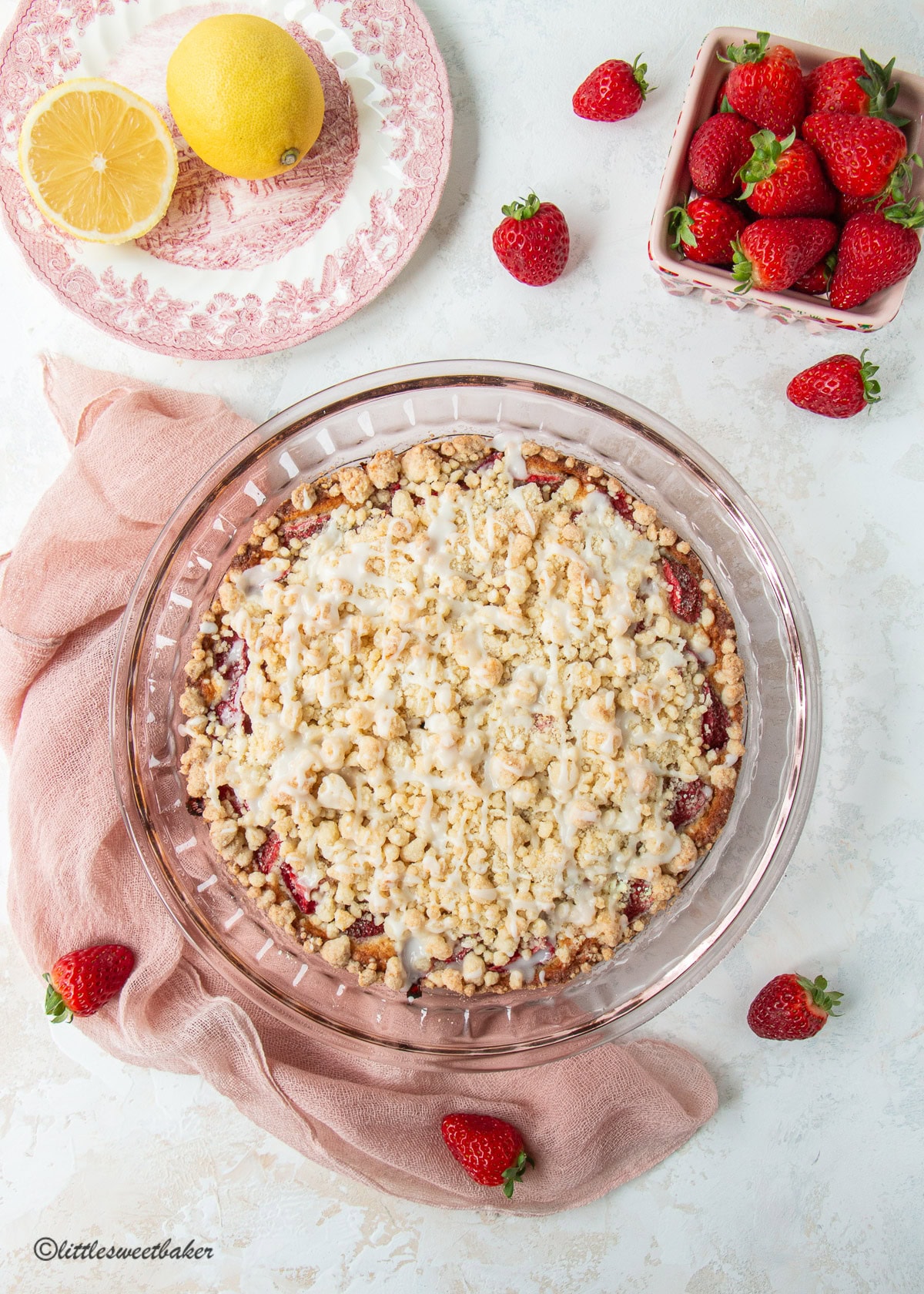 A whole strawberry crumb cake in a pink glass pie plate with strawberries, lemons, and a pink chiffon napkin.