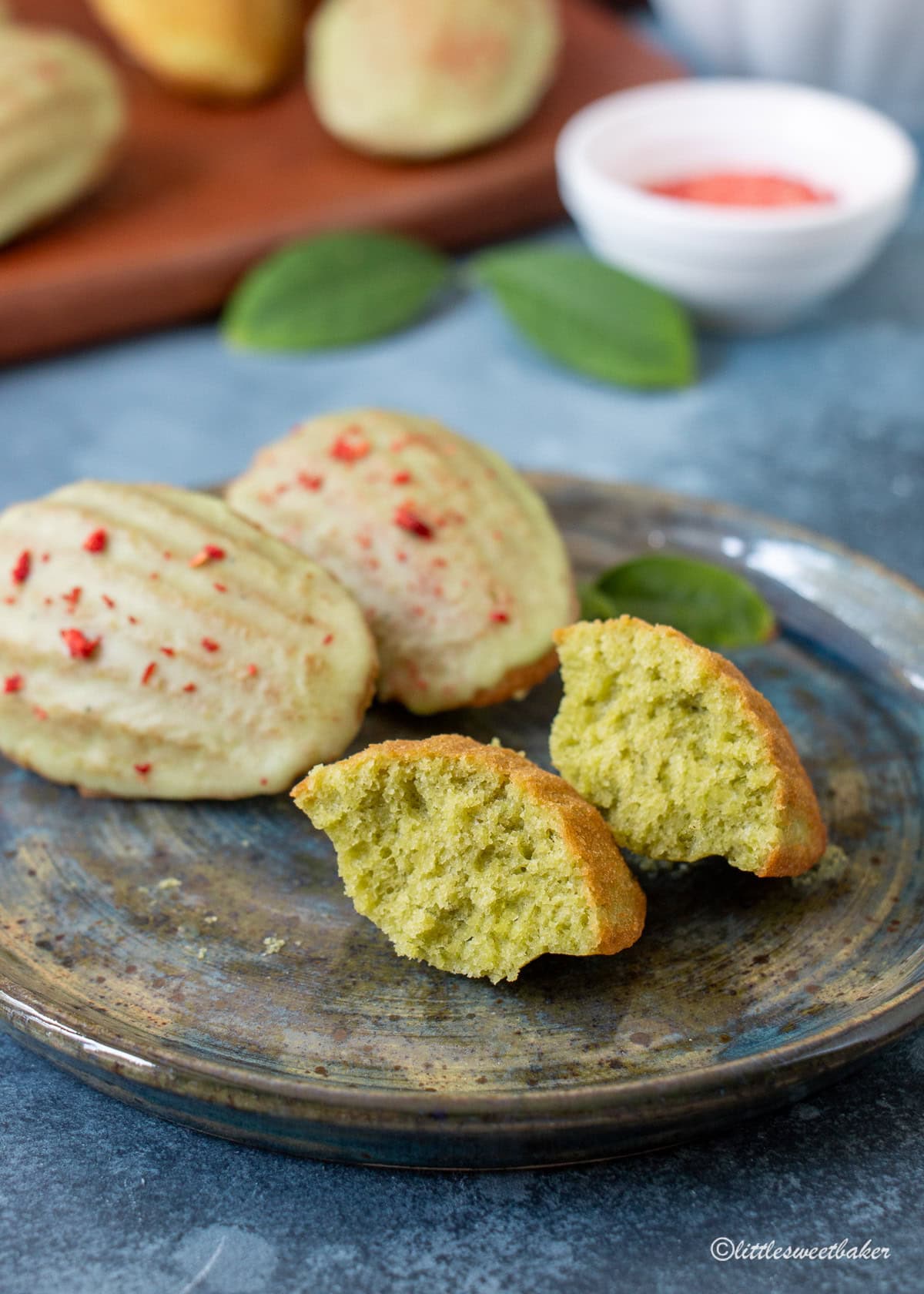 Glazed matcha madeleines topped with finely crushed freeze-dried strawberries on a plate with one broken in half.