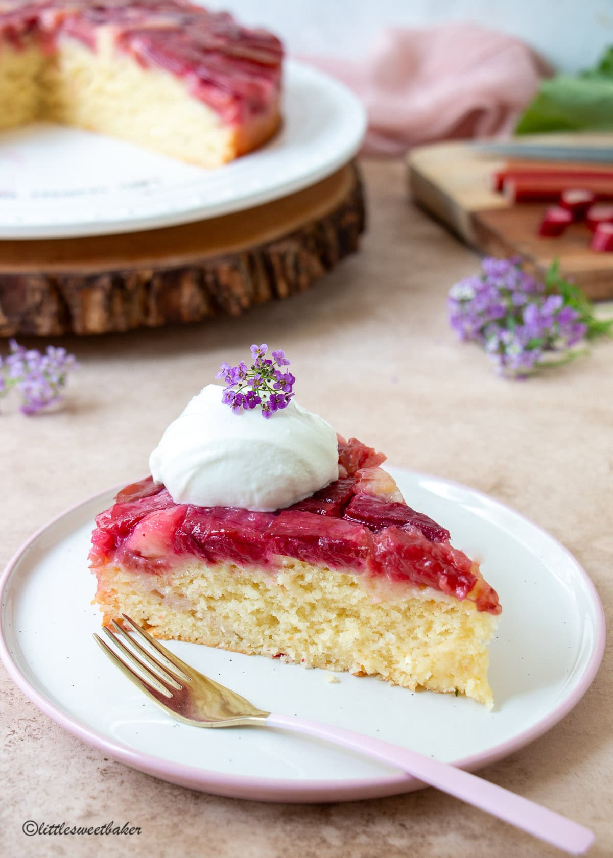 A slice of rhubarb upside-down cake topped with whipped cream and some tiny purple flowers.