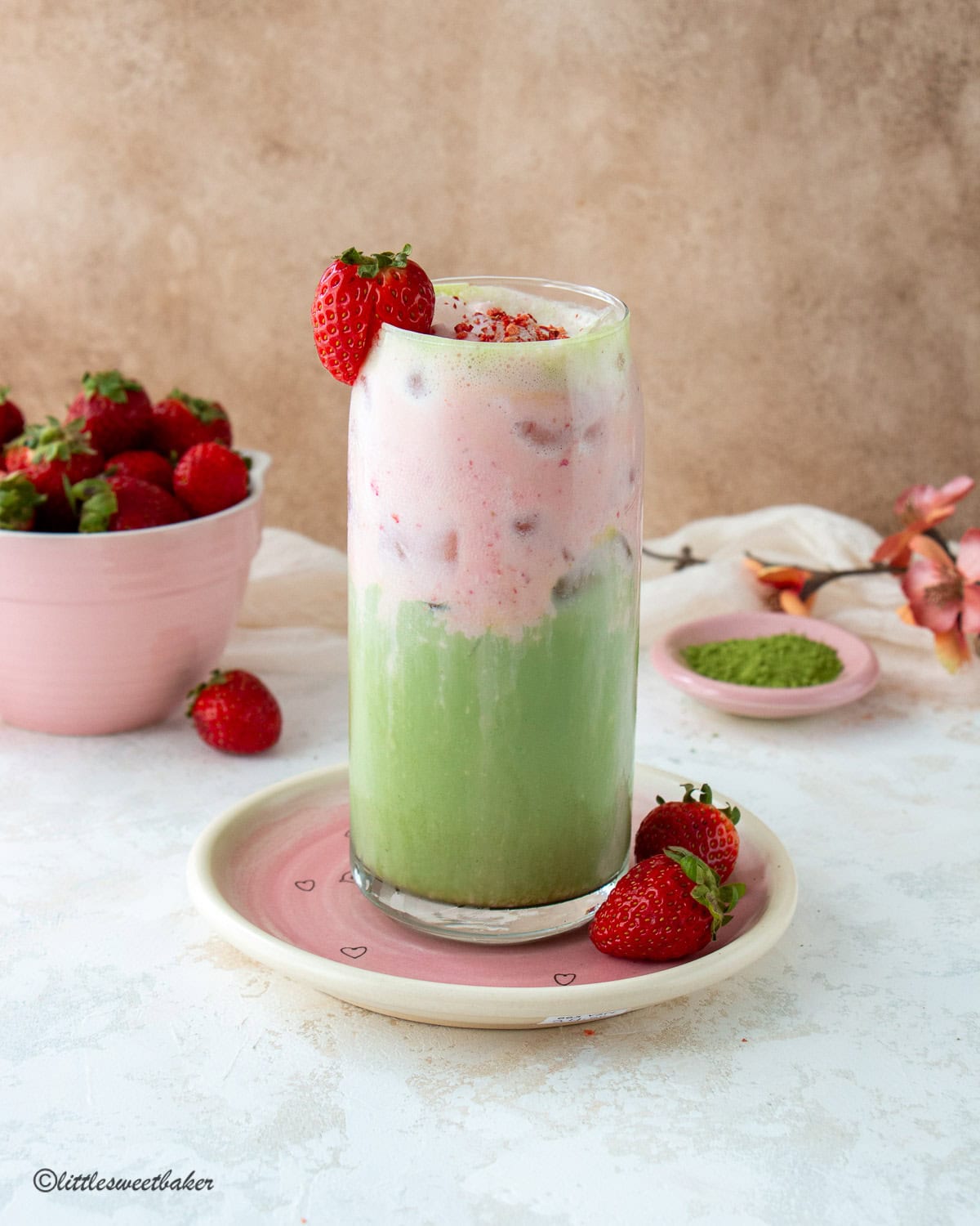 A glass of strawberry protein cold foam matcha latte on a pink plate with strawberries.