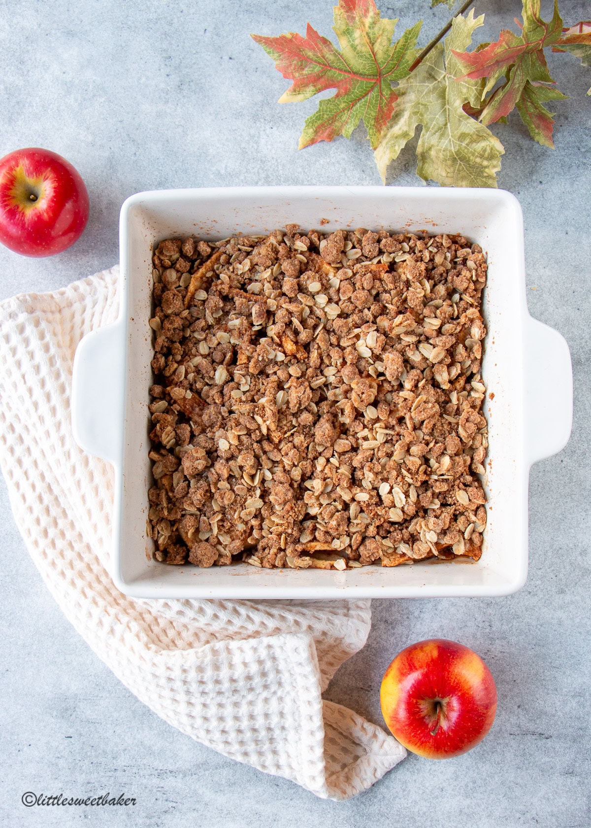 Healthy apple crisp in a white square baking dish with a with towel and two apples.