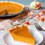 A slice of healthy pumpkin pie on a light grey plate with the rest of the pie in the background.