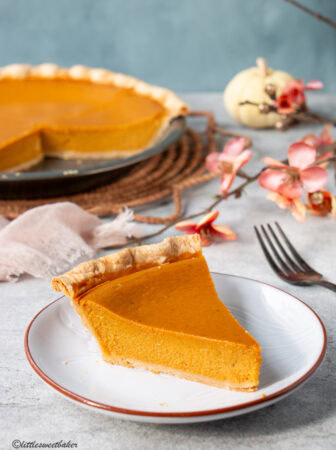 A slice of healthy pumpkin pie on a light grey plate with the rest of the pie in the background.