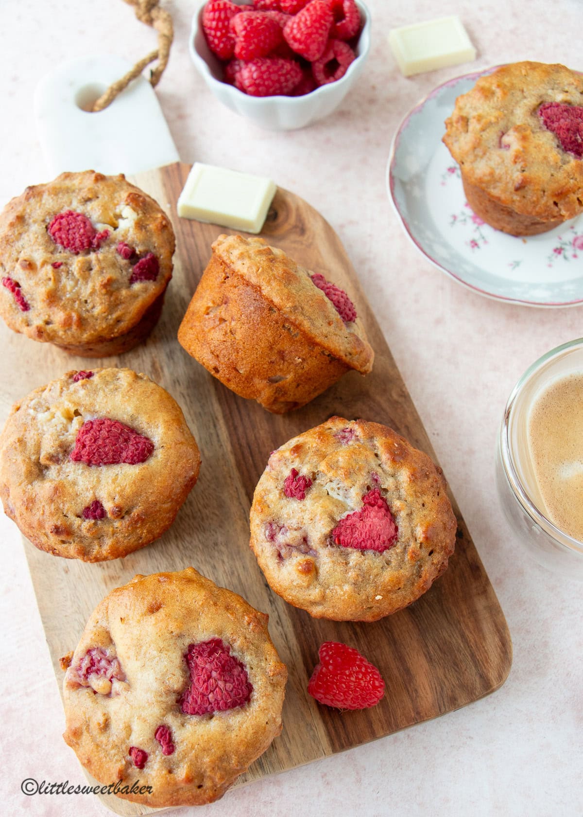 High-fiber raspberry bran muffins on a wooden cutting board surrounded by a bowl of fresh raspberries and cup of coffee.