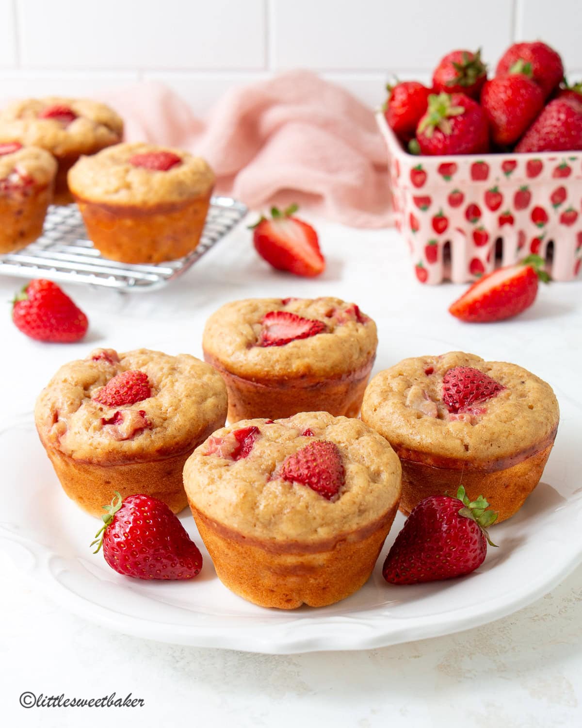 A plate with high-protein strawberry muffins with a basket of strawberries and more muffins in the background.