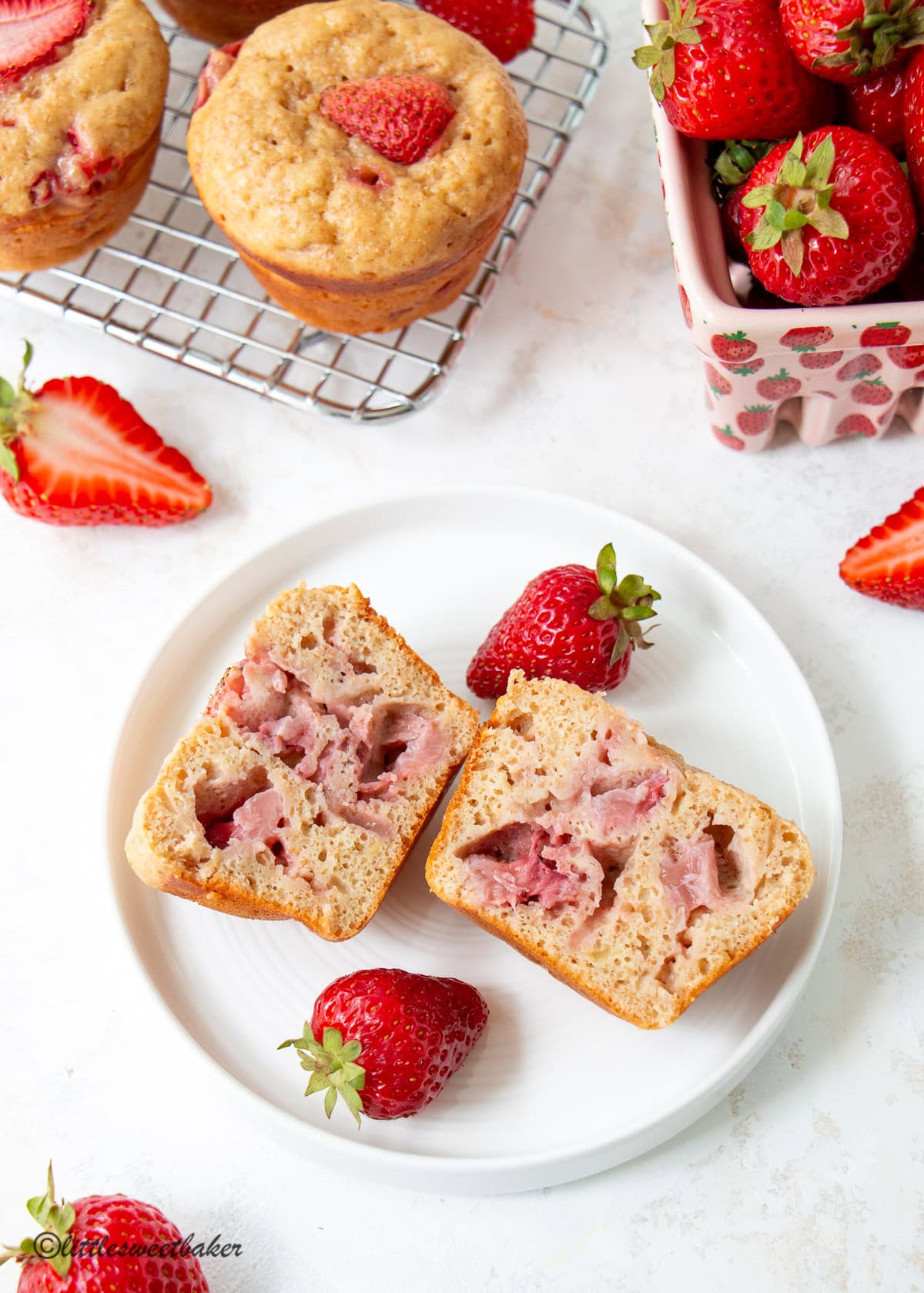 A high-protein strawberry muffin cut in half on a white plate surrounded with strawberries and more muffins.
