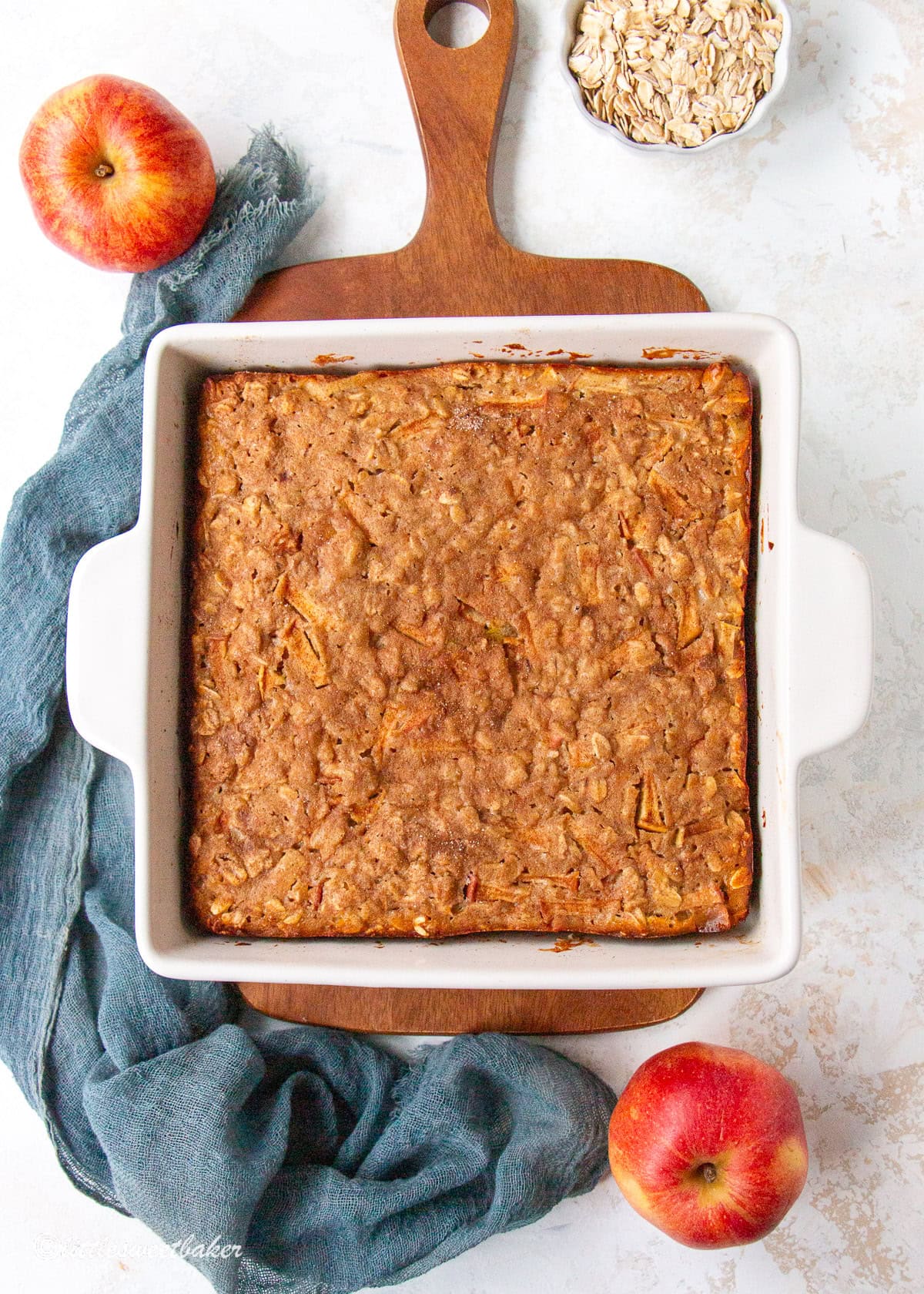 A healthy apple oatmeal cake in a white square baking pan on top of a wooden cutting board.